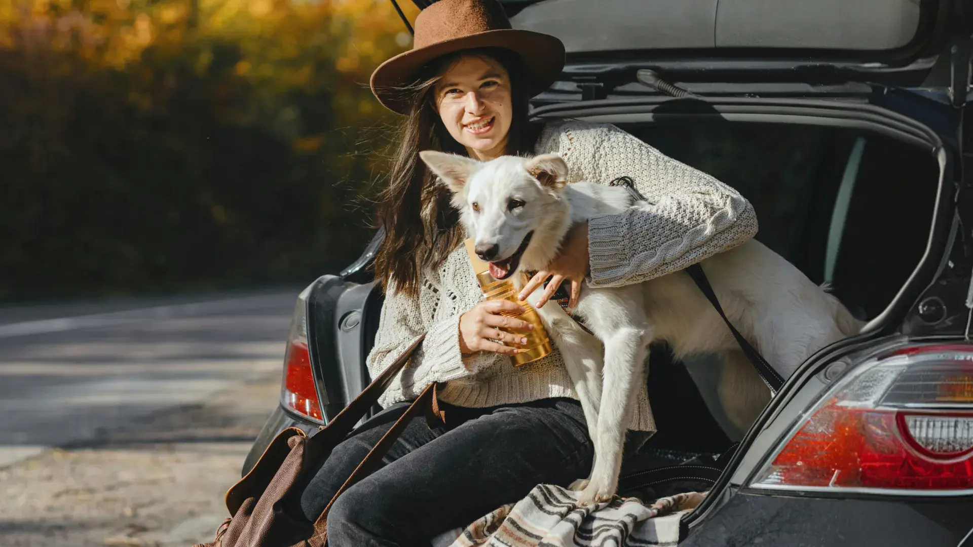 A smiling woman in a brown hat and cream sweater sits in the open trunk of a car, hugging a large, happy white dog during an autumn road trip.