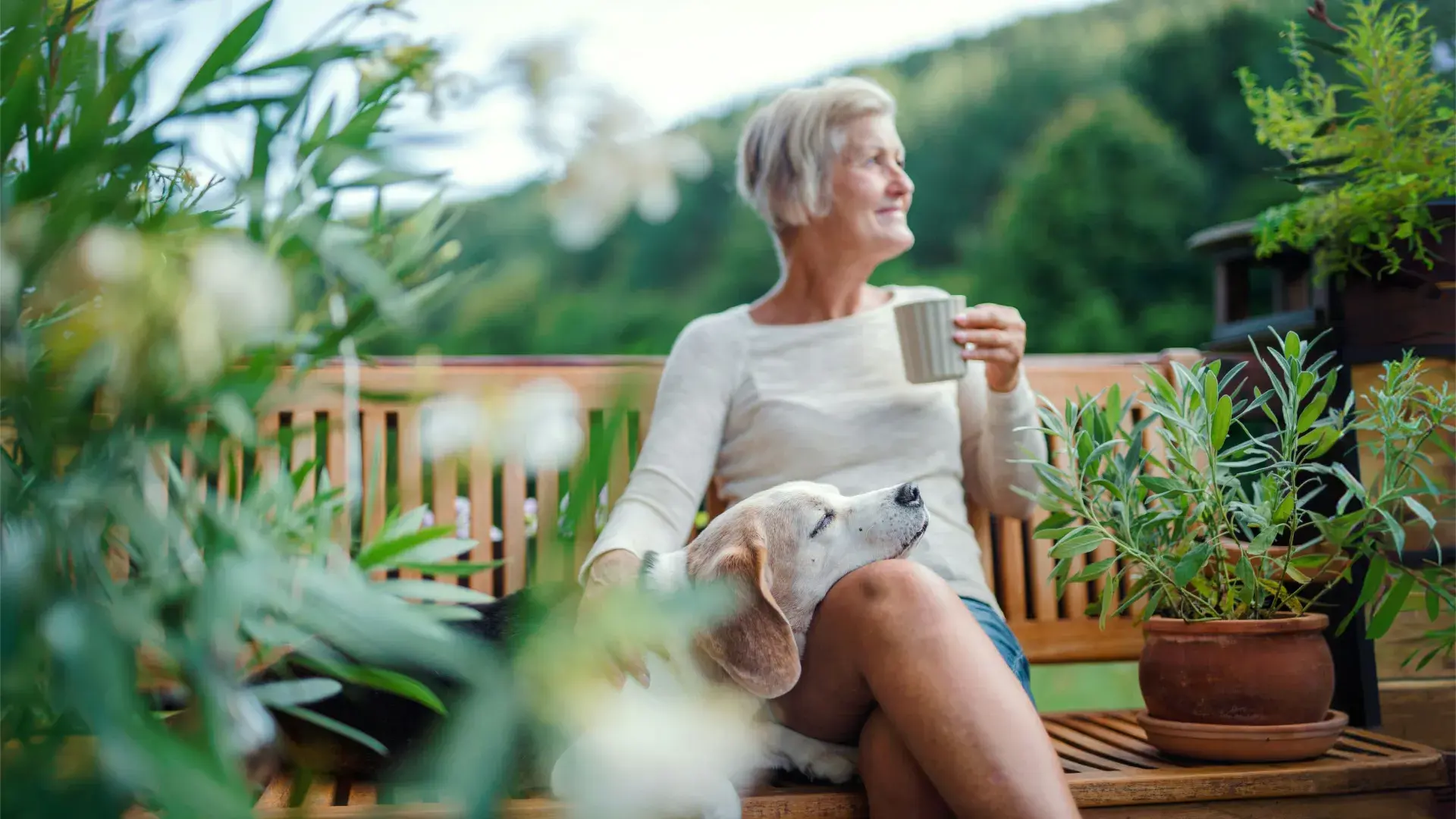 A senior woman with short blonde hair sits on a wooden garden bench, holding a mug and looking off-camera. An older beagle rests its head comfortably on her lap. The scene is set on a lush, green outdoor balcony or patio surrounded by potted plants.