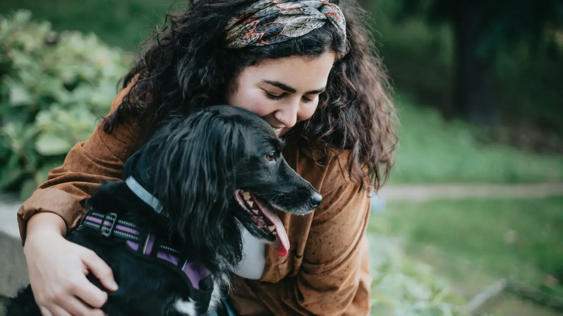 A close-up shot of a young woman leaning down to affectionately nuzzle a black dog. The dog is panting happily with its tongue out, showing a strong bond between the pet and its owner in an outdoor grassy area.