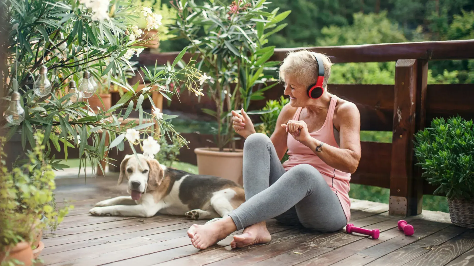 An active senior woman wearing red headphones and a pink tank top sits on a balcony deck, smiling and gesturing while her senior beagle lies calmly beside her. The setting is surrounded by lush green plants and outdoor light strings.