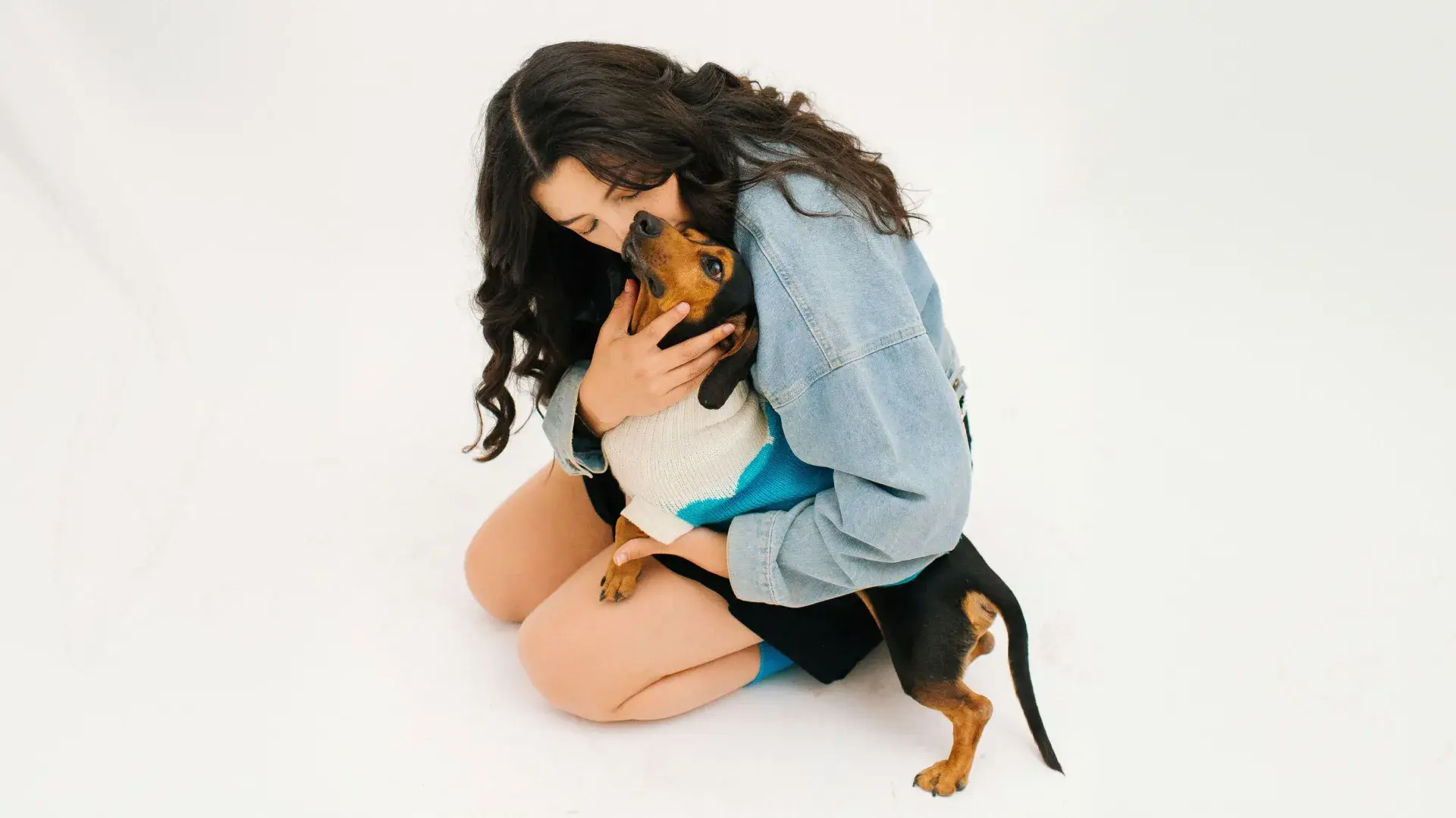 A woman in a denim jacket kneeling on a plain white background and affectionately hugging a small black and tan Dachshund wearing a blue and white sweater, symbolizing the strong emotional bond and love between humans and their pets.