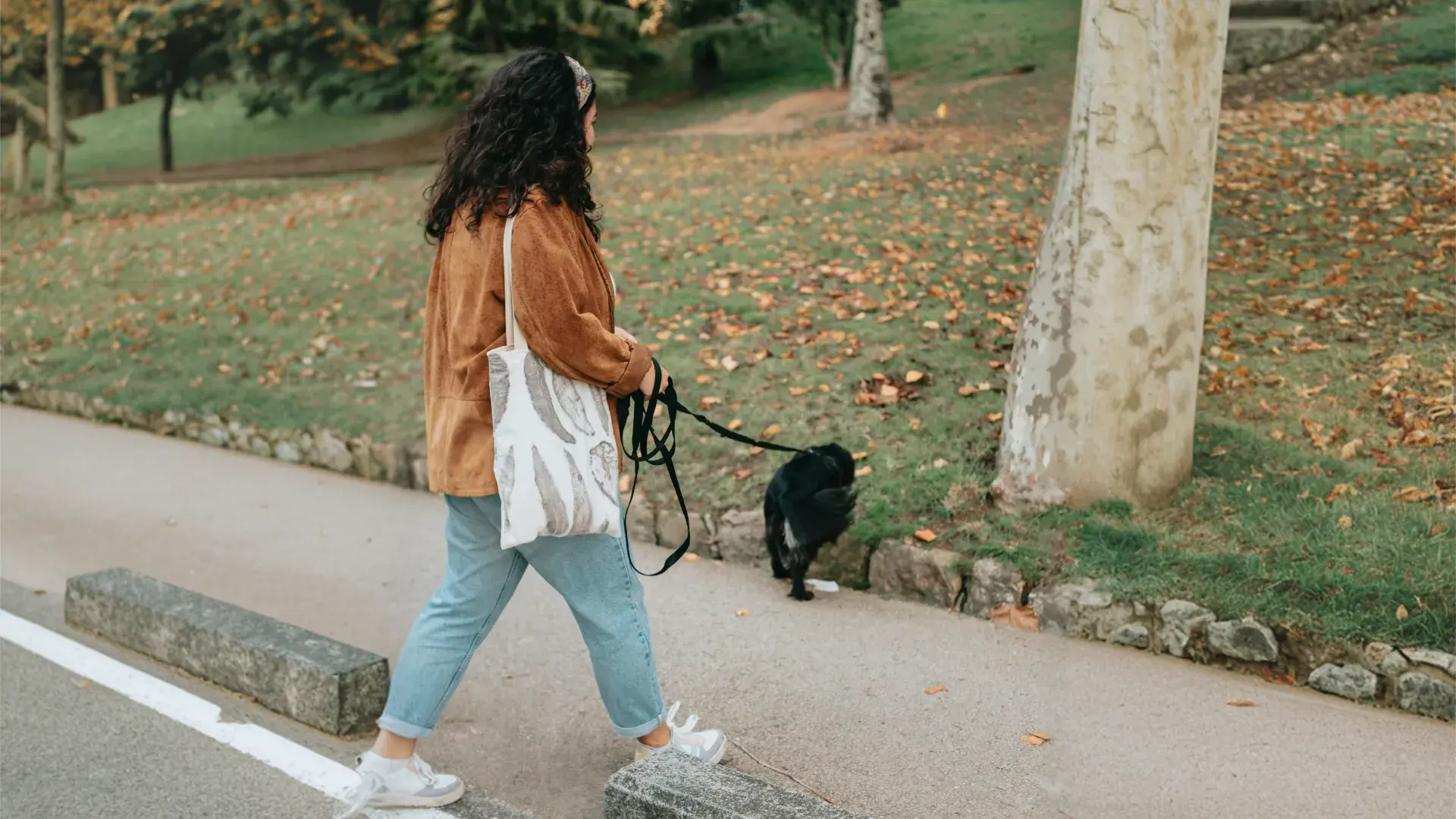 A woman in a brown jacket and blue jeans walks her small black dog on a leash along a paved path in a park. She is carrying a white tote bag, capturing a typical daily bonding activity.