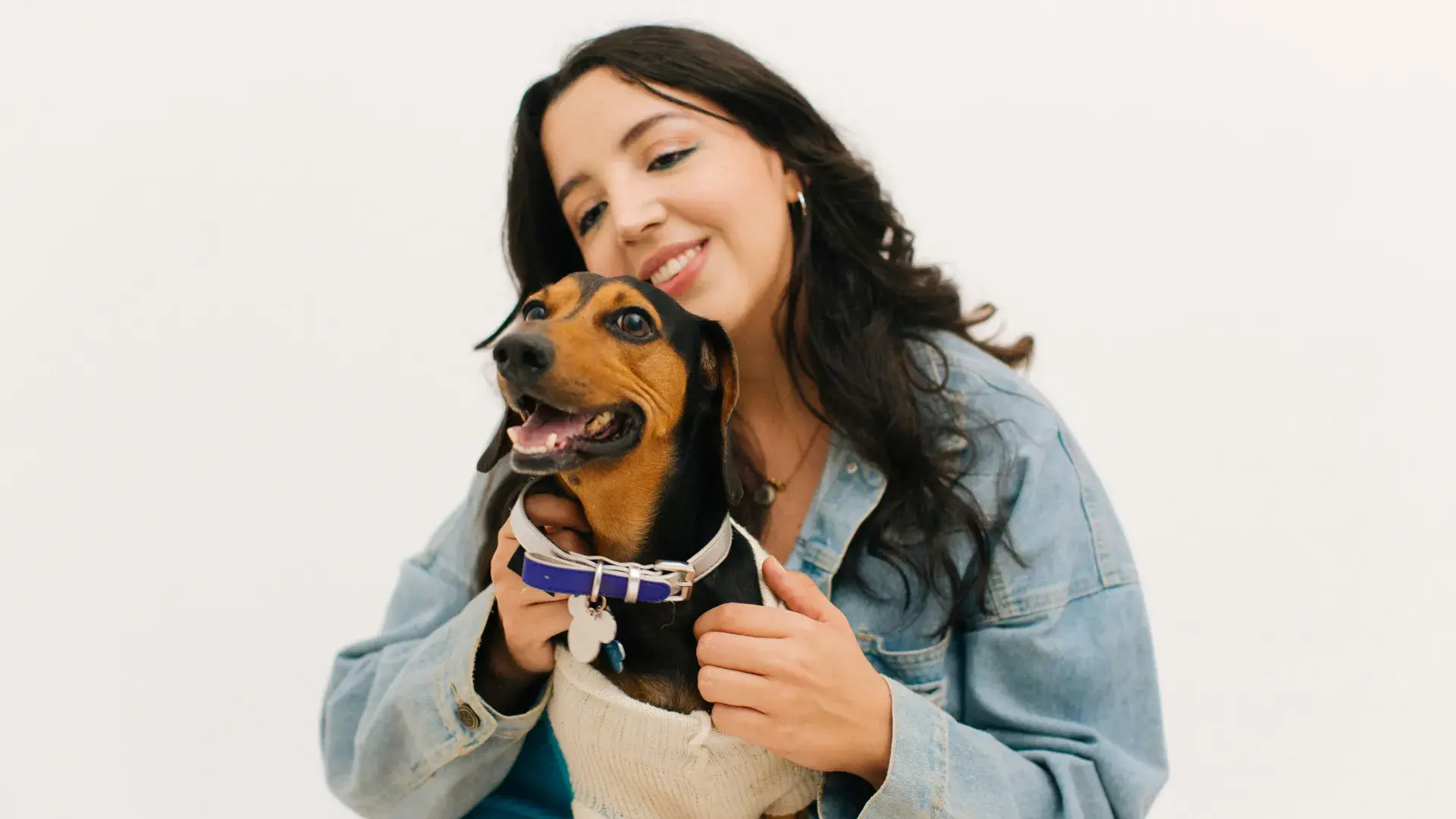 A woman in a denim jacket tenderly hugging a small black and tan Dachshund in a blue sweater against a white background, illustrating the deep emotional connection between dogs and humans.