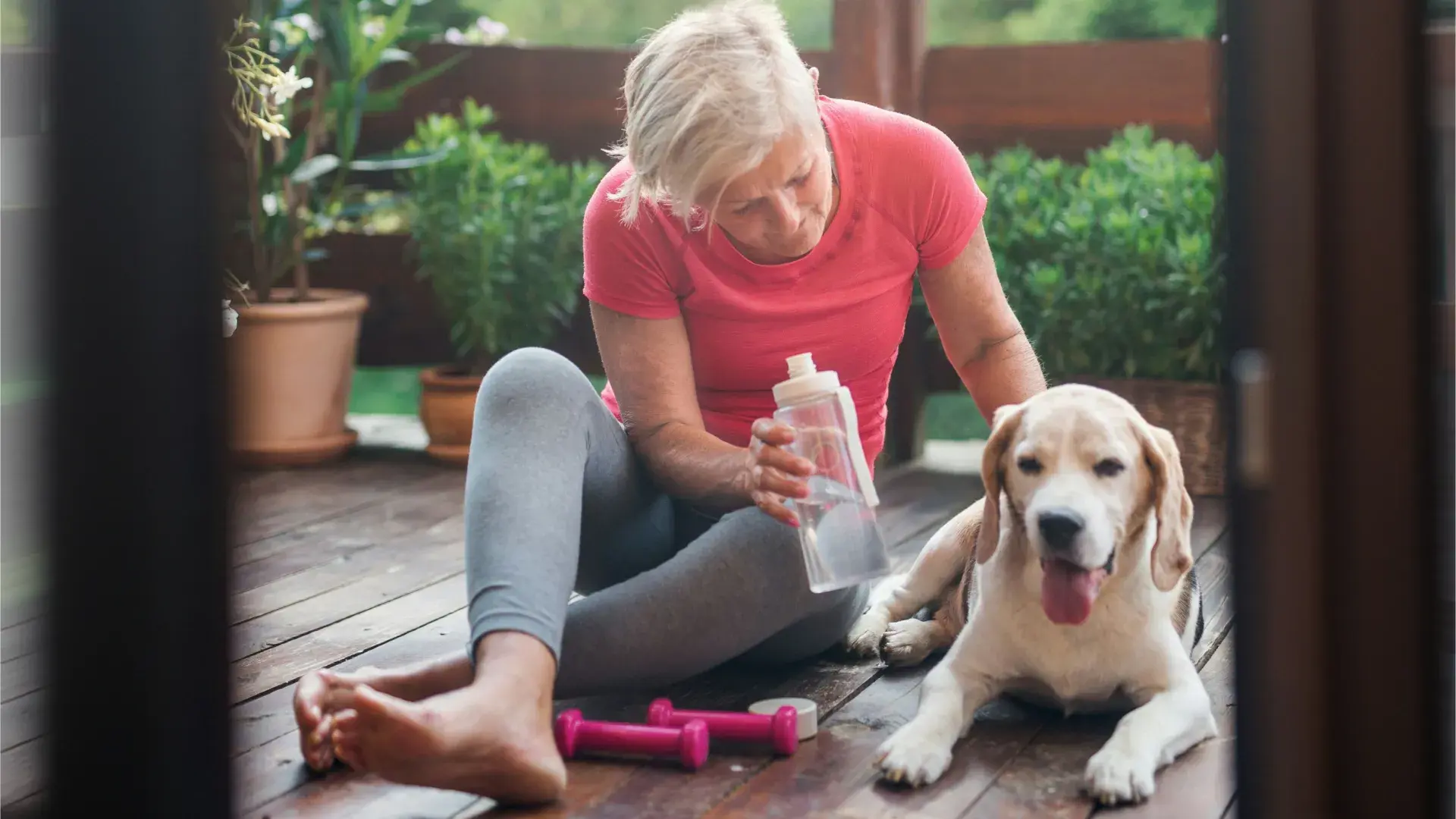 A senior woman in a pink workout shirt sits on a wooden deck, holding a water bottle and looking down at her senior beagle. Small pink dumbbells lie on the floor nearby, suggesting a break during a light exercise or training session.
