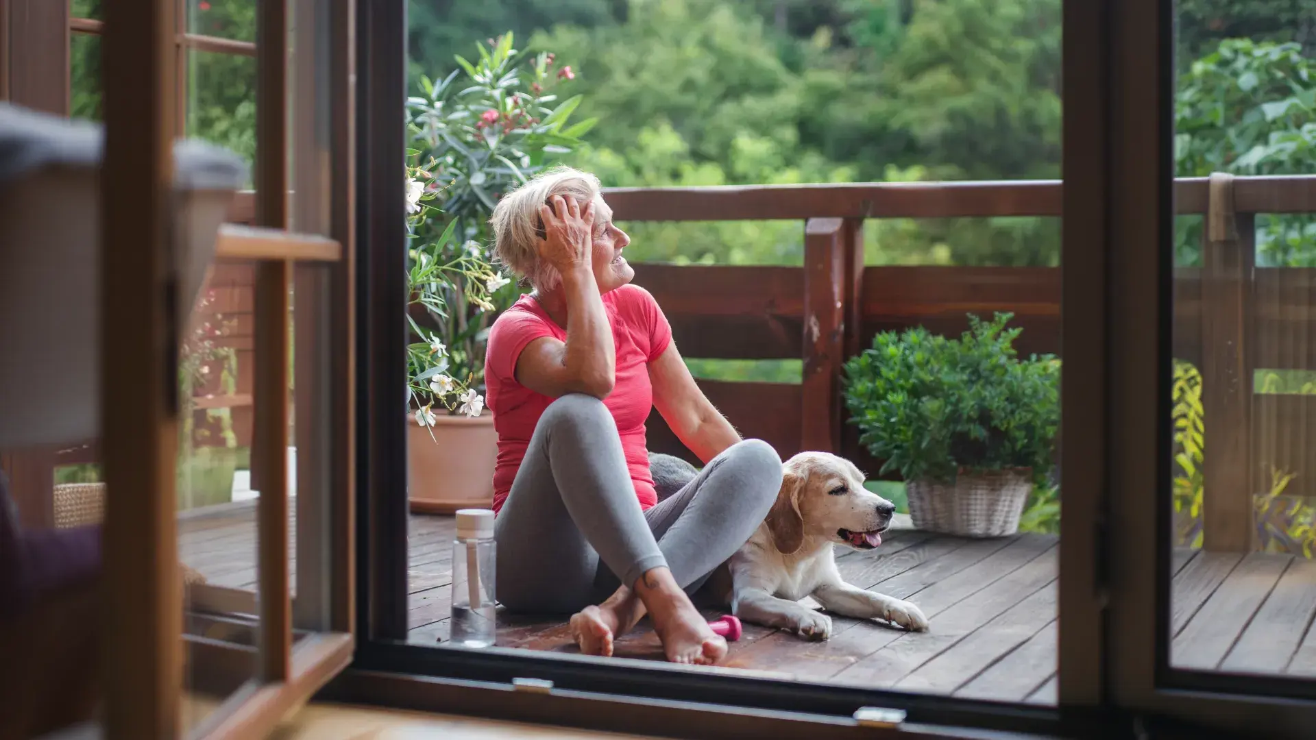 A senior woman in a pink top and grey leggings sits happily on a wooden deck next to her beagle. She runs a hand through her short blonde hair while looking out at the lush greenery. A water bottle is on the floor.